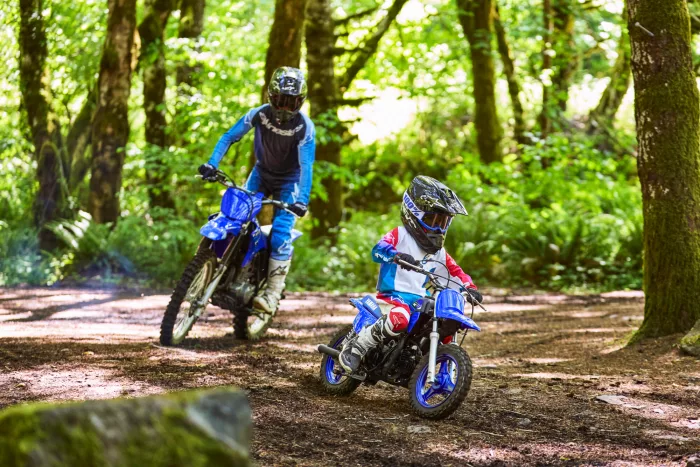 Two young motocross riders in helmets navigate a dirt trail through a sunlit forest.