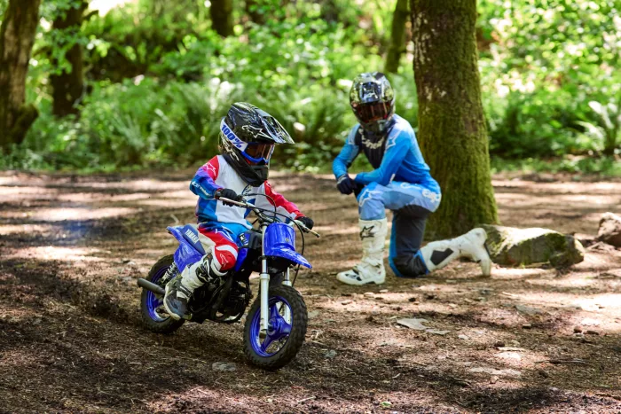 Two young riders wearing full protective gear ride dirt bikes through a sunlit forest trail, one in front on a blue bike and another behind in blue gear.