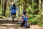 Two dirt bike riders wearing helmets navigate a rocky forest trail, foreground rider on a blue bike in blue gear.