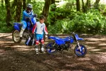 Two kids in motocross gear beside blue dirt bikes in a forest clearing.