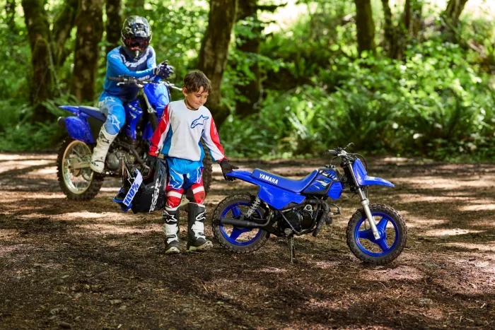 Two kids in motocross gear beside blue dirt bikes in a forest clearing.