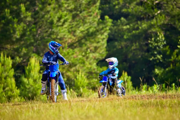 Two motocross riders in blue gear on blue dirt bikes riding through a grassy field with trees in the background.