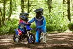 Adult and child in full motocross gear with helmets beside a blue dirt bike on a forest trail.