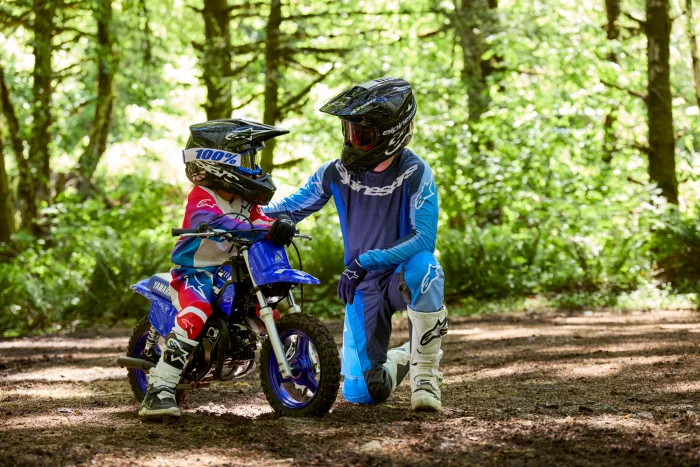 Adult and child in full motocross gear with helmets beside a blue dirt bike on a forest trail.