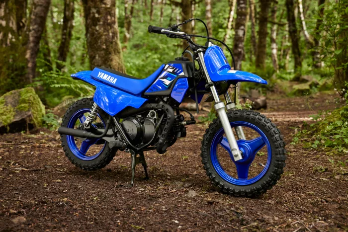 Blue Yamaha dirt bike resting on its kickstand in a forest clearing, with knobby tires visible in the dirt trail.