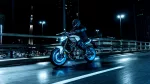 Motorcycle rider in full protective gear on a white-and-blue MT-09 bike, riding on a wet city bridge at night with bright streetlights in the background.