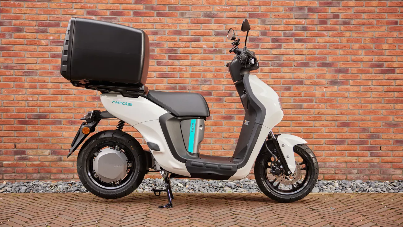White Yamaha scooter with a large black top case attached, parked against a brick wall backdrop.