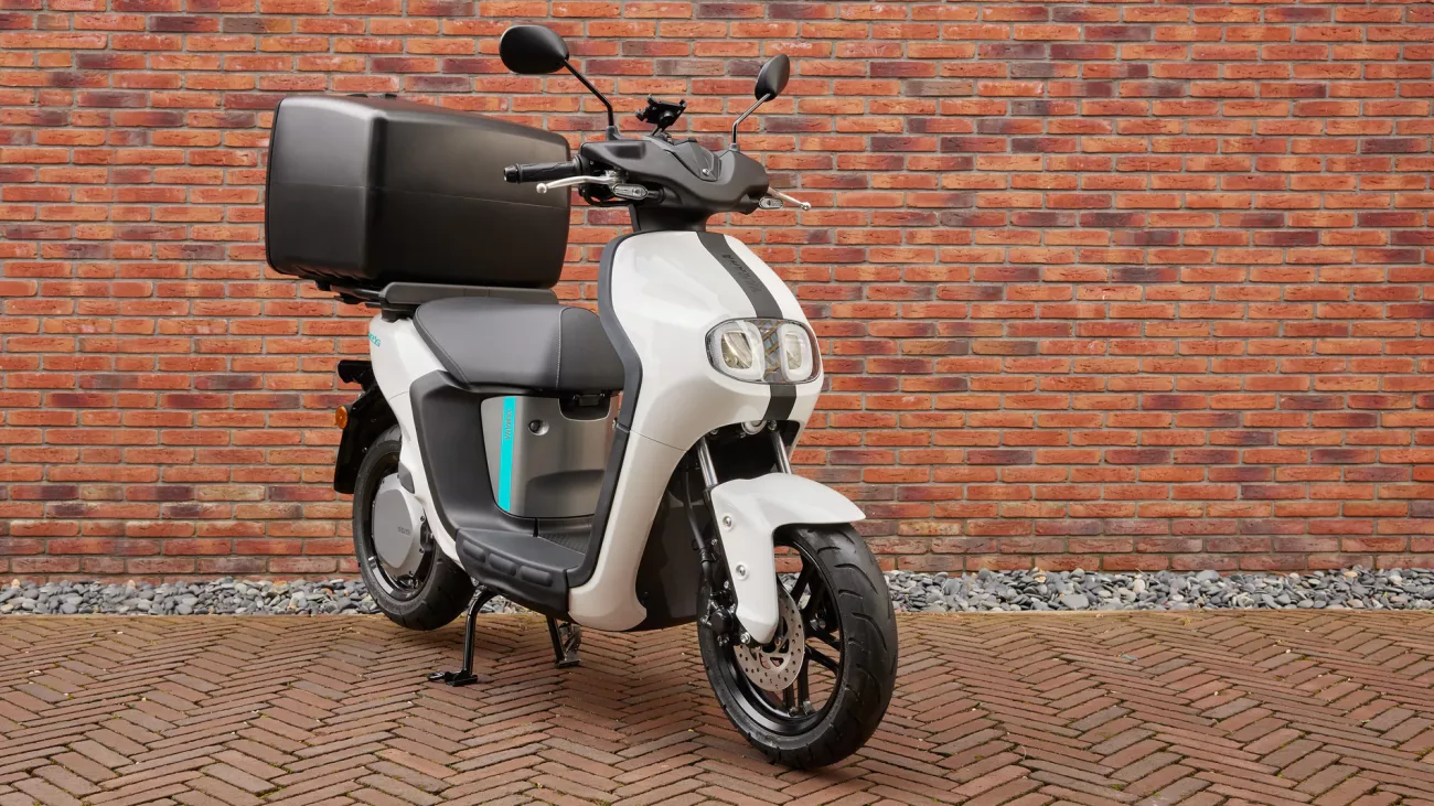 White scooter with a large black top case parked on a brick-paved surface in front of a red brick wall.