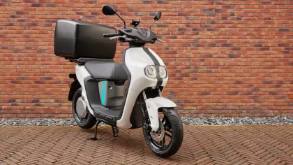 White scooter with a large black top case parked on a brick-paved surface in front of a red brick wall.